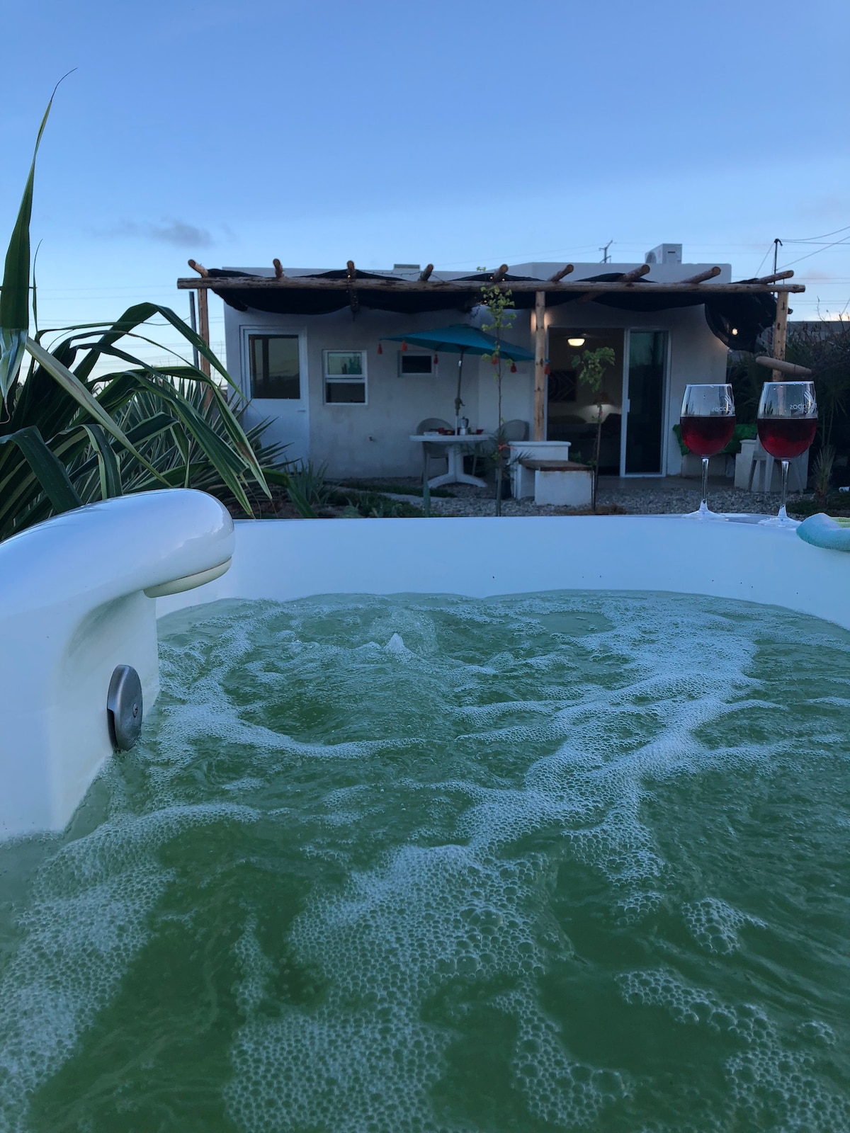 A spacious jacuzzi is shown in the foreground, filled with bubbling water. Two glasses of red drink are placed nearby. In the background, a casita with a shaded patio and a garden area is visible, framed by lush green plants.