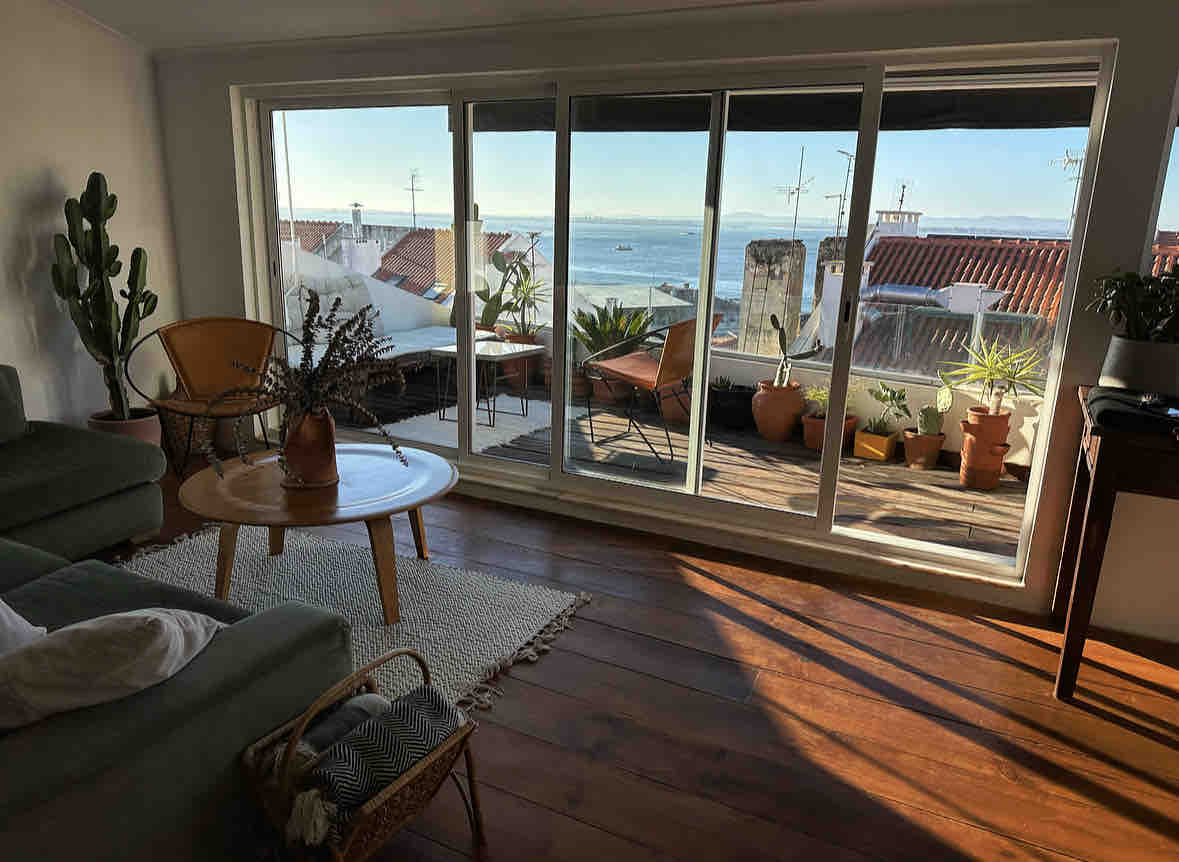 A sunlit living space features large glass doors opening to a terrace adorned with various potted plants. A low wooden table sits on a textured rug, while a comfortable sofa adds to the inviting atmosphere. A captivating view of the water completes the scene.