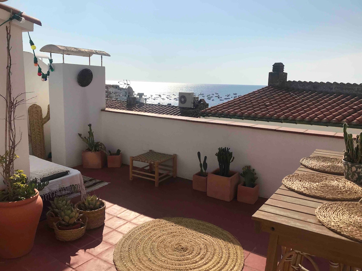 A sunlit terrace features a rustic wooden dining table surrounded by potted plants and decorative items. The view captures the glistening sea and distant boats, with rooftop tiles in the background. A cozy seating area is positioned beside a low table, creating a relaxing atmosphere.