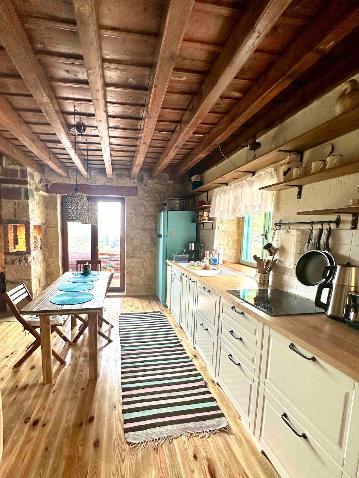 The kitchen features a rustic design with wooden beams and stone walls. A long table is set with blue plates, and a striped rug runs across the floor. Natural light enters through windows, highlighting the kitchen appliances and shelving.