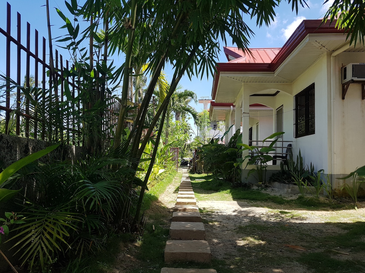 A stone pathway lined with tropical greenery leads to a cluster of simple buildings. The path is flanked by vibrant palm leaves and shrubs, creating a tranquil and inviting atmosphere. Clear blue skies and bright sunlight illuminate the serene surroundings.