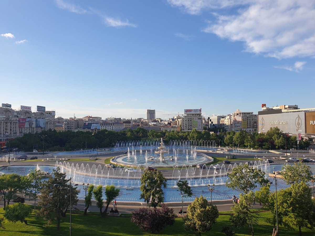 A panoramic view of Unirii Square showcases a large fountain surrounded by vibrant greenery and trees. The water displays are arranged in a circular pattern, with fountains dancing gracefully. The skyline features buildings and advertisements in the background, with a clear blue sky overhead.