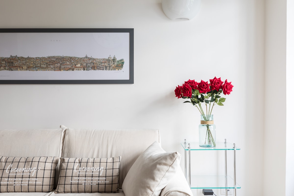 A light-filled living area features a beige sofa adorned with patterned cushions, complemented by a nearby glass table holding a vase of vibrant red roses. A panoramic framed artwork depicting the cityscape is elegantly displayed above the sofa.