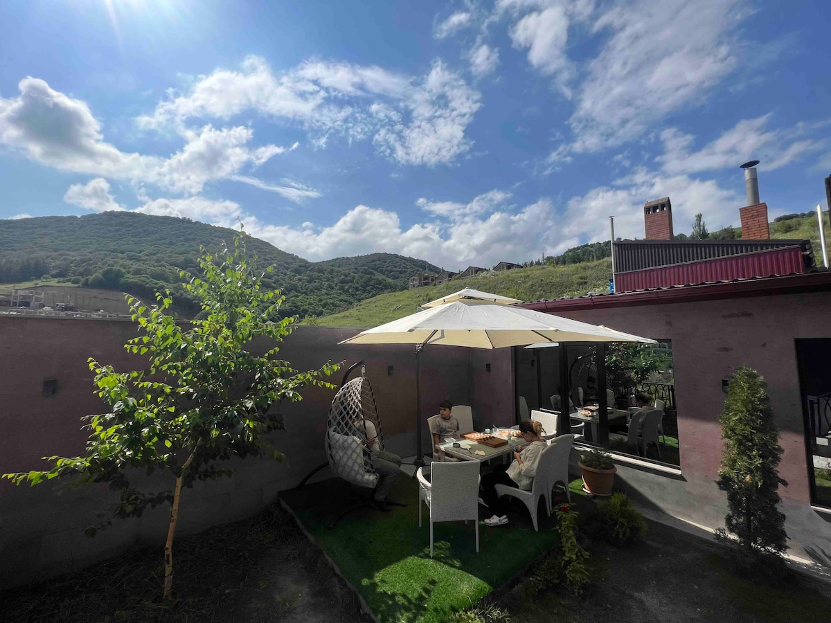 An outdoor seating area is surrounded by greenery, featuring a large umbrella providing shade over a table set for dining. A hanging chair is positioned nearby, with mountainous scenery visible in the background under a bright, blue sky.