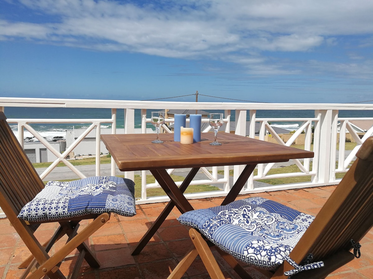 A wooden table is set on a sunlit deck, accompanied by two folding chairs adorned with blue and white cushions. A candle and glasses are placed on the table, with expansive sea views visible beyond the railing.