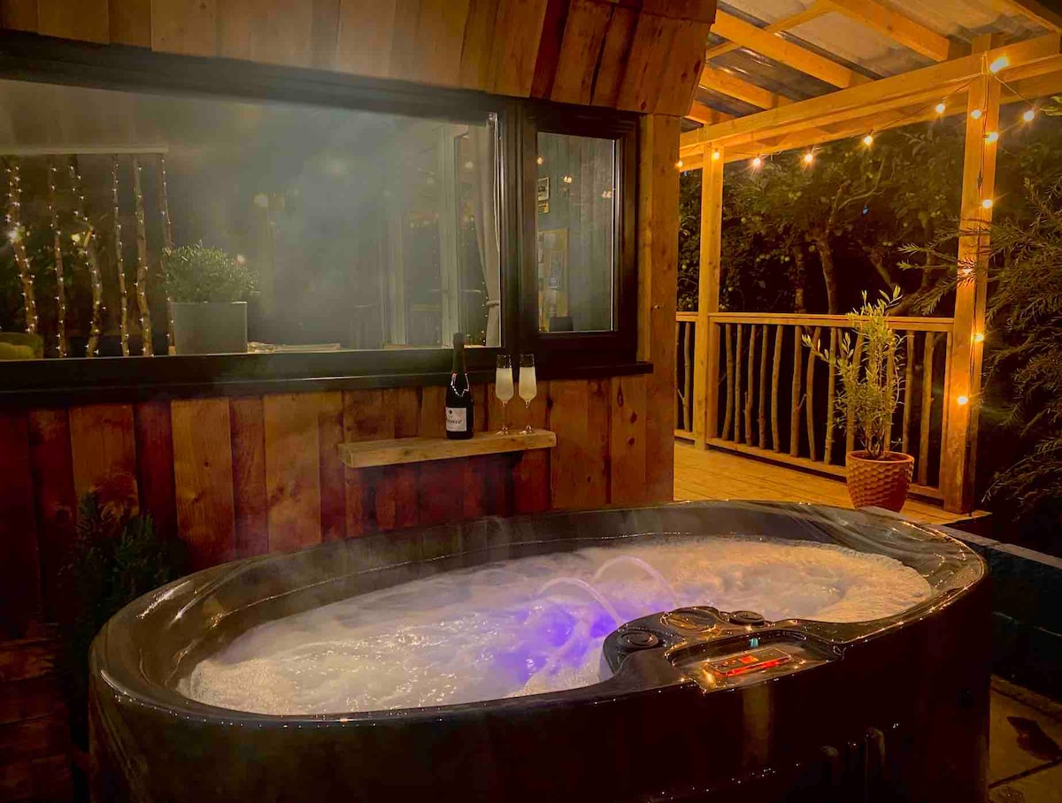 A hot tub spa is seen, set against a backdrop of wooden paneling and ambient lighting. Bubbles rise from the water, illuminated by soft, colorful lights. Two champagne flutes and a bottle of sparkling wine rest on a small shelf nearby, enhancing the inviting atmosphere.