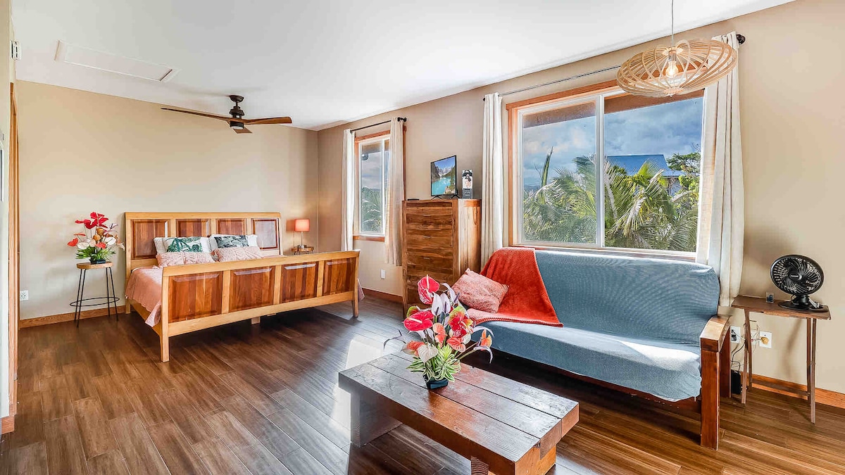The spacious bedroom features a king-sized bed against a wooden backdrop. A cozy futon couch is positioned beside a wooden coffee table adorned with tropical flowers. Large windows allow natural light to fill the room, showcasing views of surrounding greenery.