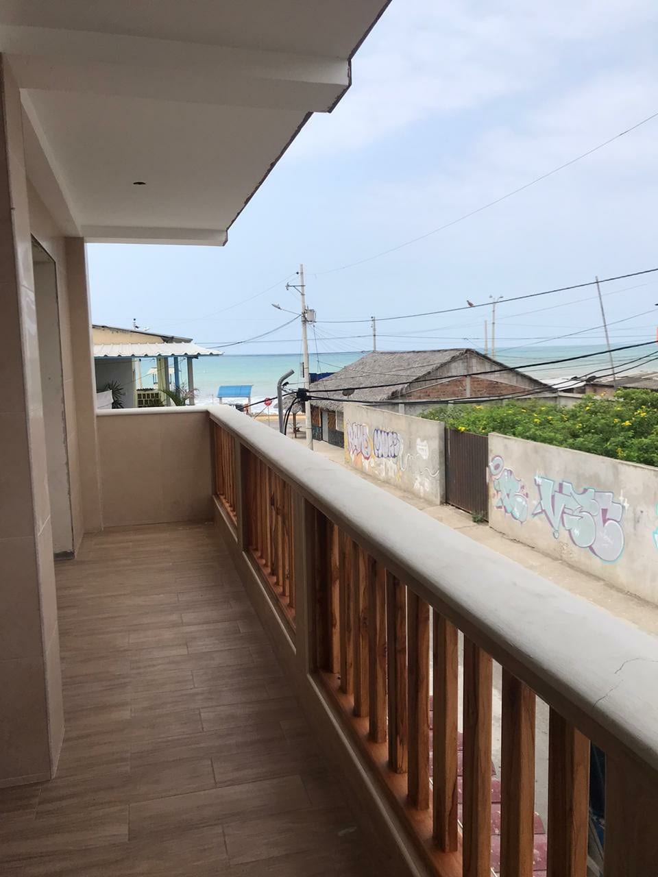 A spacious balcony features wooden railings, offering views of the ocean beyond. The surrounding area includes simple structures and electrical lines, with yellow flowers seen in the foreground. The surface is tiled, providing a clean and open space for relaxation.