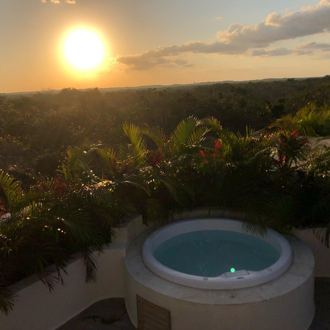 A private rooftop jacuzzi is framed by lush green foliage, offering a tranquil setting. The sun sets in the background, casting a warm glow over the distant landscape. Gentle hills and treetops are visible, creating a serene atmosphere.