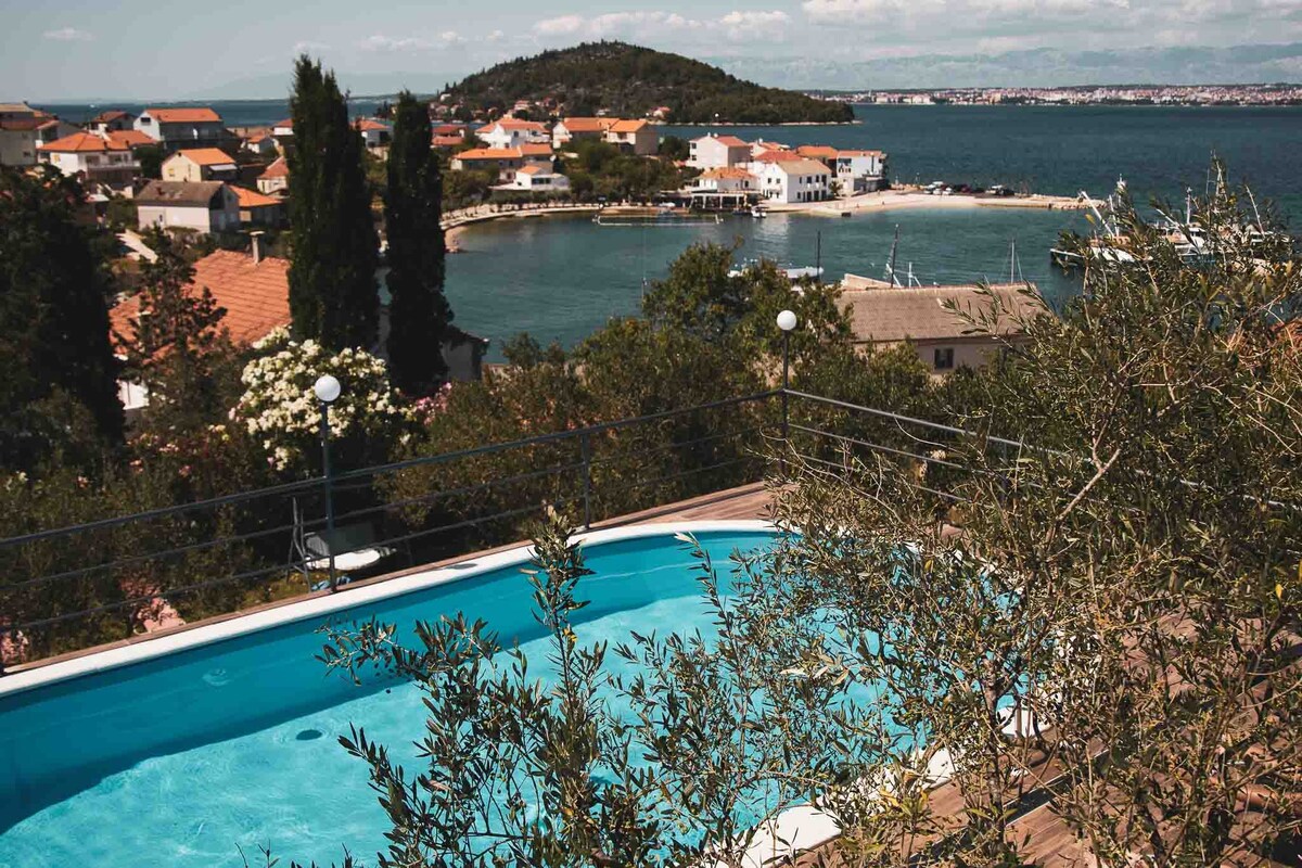 A tranquil swimming pool with a crystal-clear surface is surrounded by lush greenery, offering a serene view of the coastline. The vibrant rooftops of nearby houses and a distant island create a picturesque backdrop against a clear blue sky.