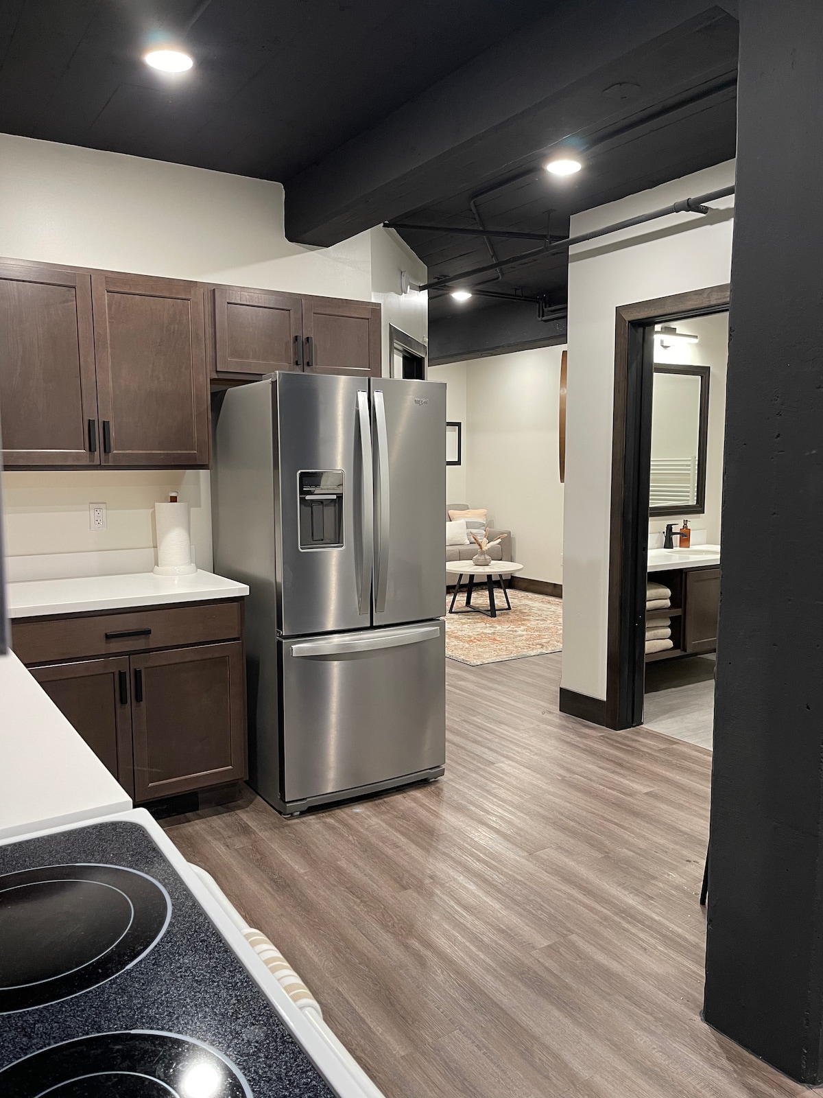The kitchen area features dark wood cabinetry and a modern stainless steel refrigerator. A stove is positioned on the countertop next to a clean white sink. The open layout leads to a living space with a small rug and seating area, highlighted by warm overhead lighting.