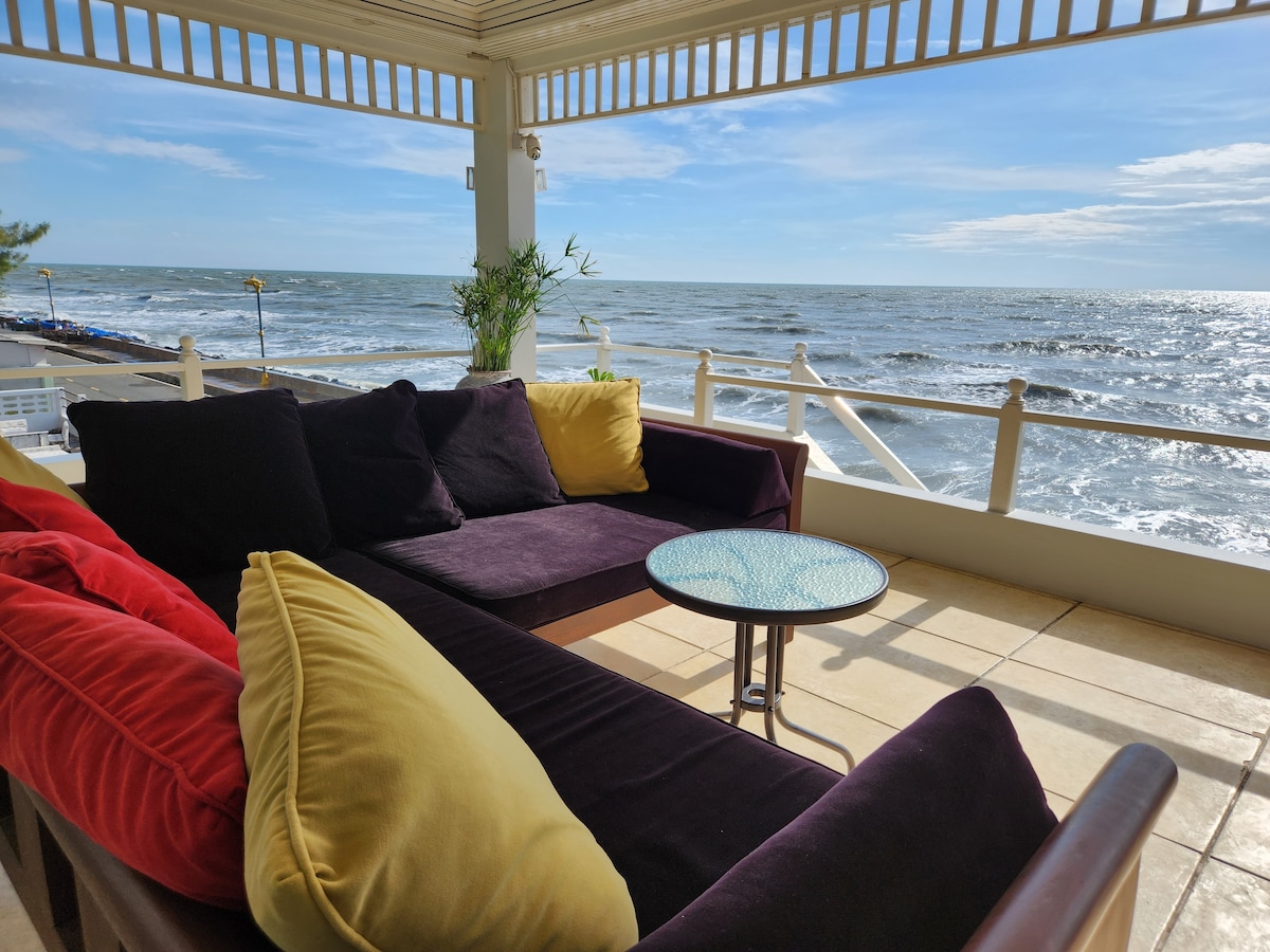 A comfortable seating area features plush cushions in deep purple and yellow, arranged on a wooden frame. A small circular table sits in front, with the ocean visible in the background. Sunlight reflects off the water, creating a serene coastal atmosphere.