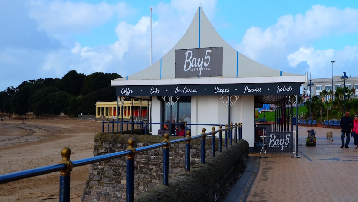 A beachfront café structure with a pointed roof displays the name 'Bay5.' Outdoor seating is available along a promenade, with a variety of food options including coffee, ice cream, and salads. The sandy beach and coastal greenery are visible in the background.