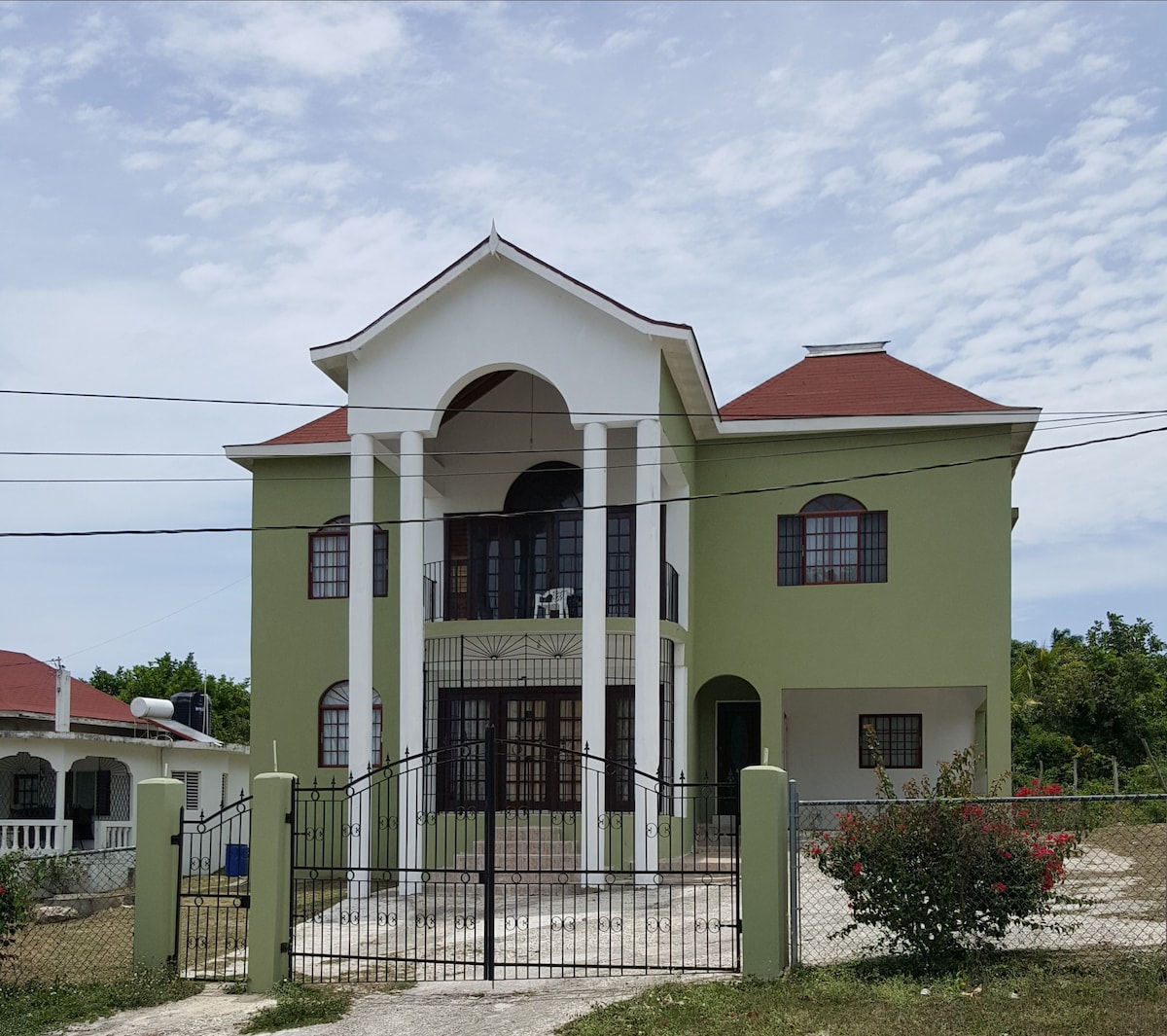 A two-story house is presented with a green exterior, featuring multiple large windows and a white columned entrance. A decorative wrought iron gate frames the property, while the surrounding landscape includes a mix of greenery and gravel pathways.