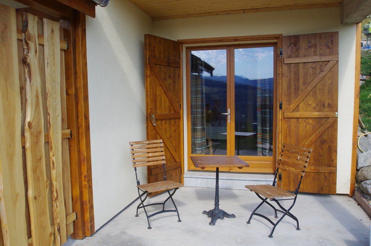 A welcoming outdoor seating area features a small table and two wooden chairs. Double doors, crafted from wood, open to reveal scenic views beyond, with light drapes partially visible inside. The entrance is complemented by stone and wooden elements, creating an inviting atmosphere.