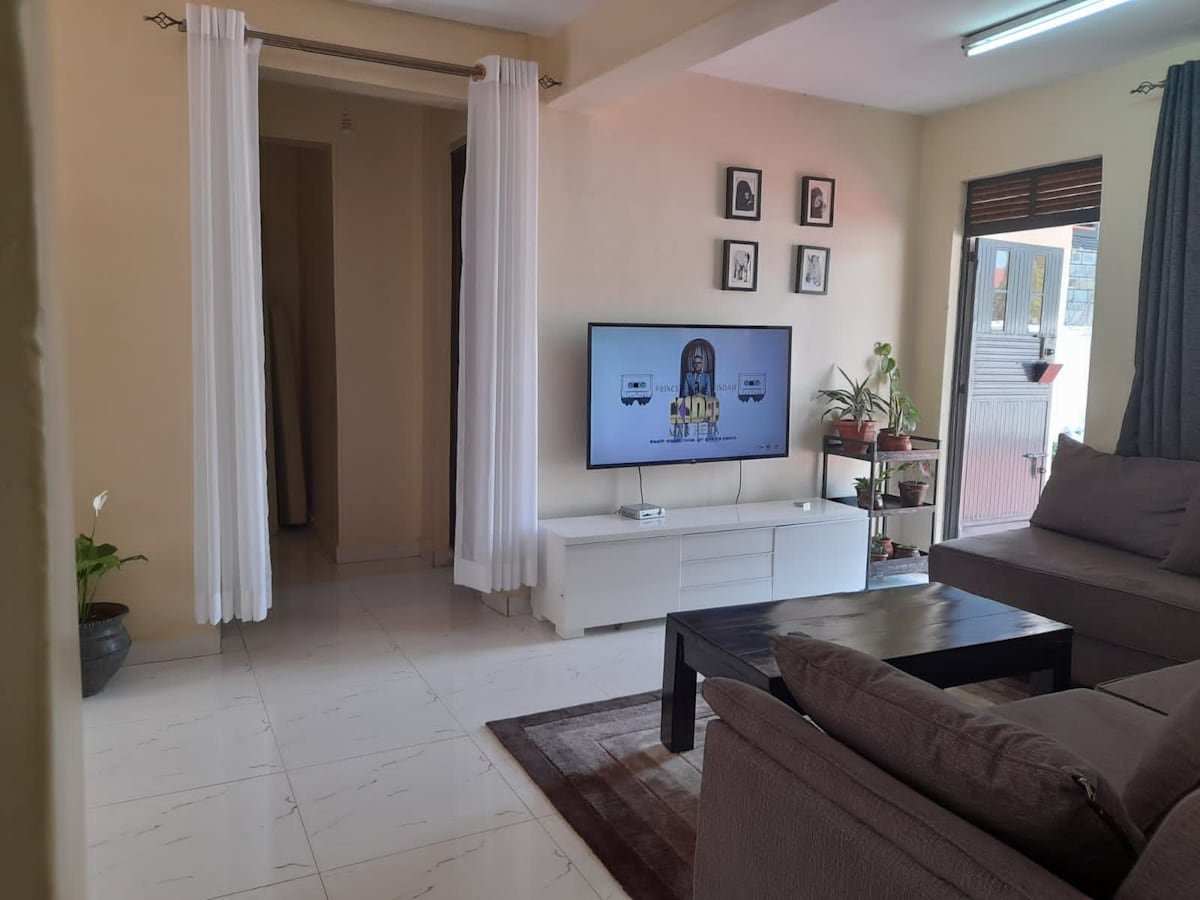A bright living area is displayed, featuring a large flat-screen television mounted on a white media console. A comfortable gray sectional sofa and a dark coffee table are positioned on a spacious tiled floor. Light-colored curtains frame the window, complementing the warm wall tones.