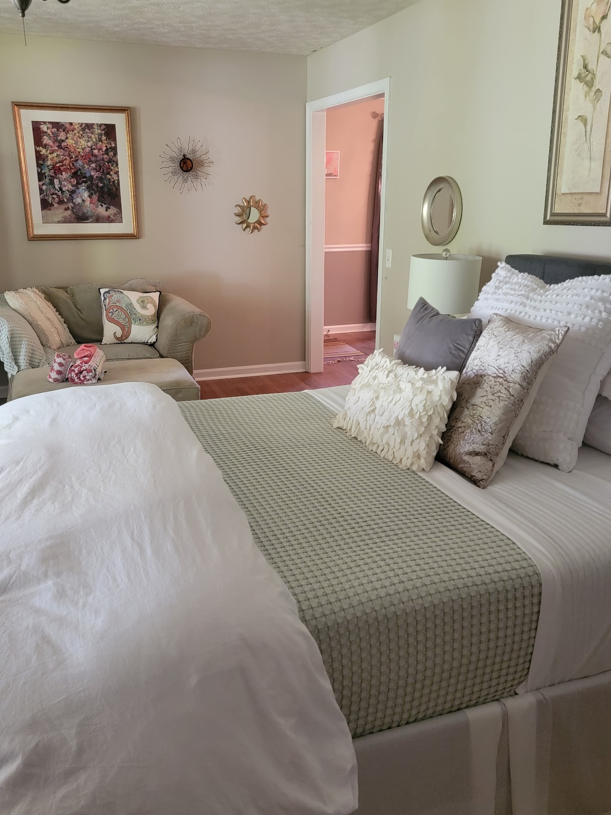 A serene bedroom space features a spacious bed adorned with patterned and textured pillows. A comfortable chair is positioned nearby, complemented by soft decorative accents and artwork on the walls. Light natural tones create a calming atmosphere, inviting relaxation.
