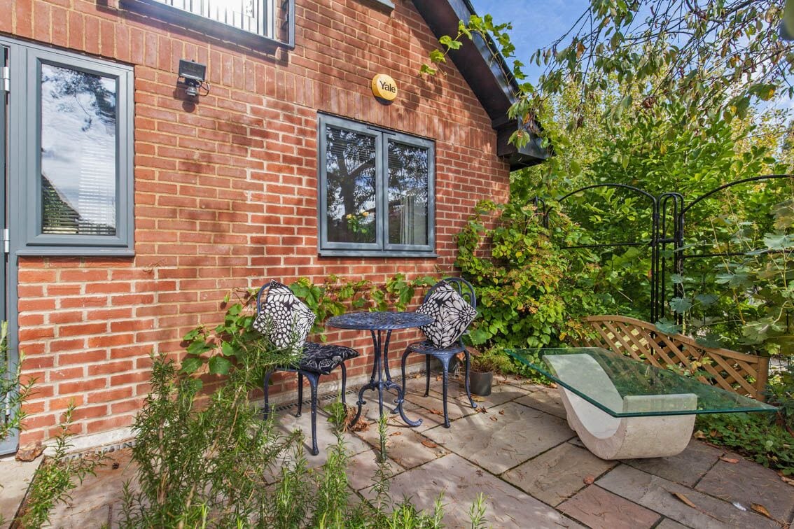 A charming outdoor seating area features a round table surrounded by two patterned chairs. Lush greenery and climbing plants create a private atmosphere, while a glass-top coffee table is placed on the stone patio. Natural light enhances the inviting setting.