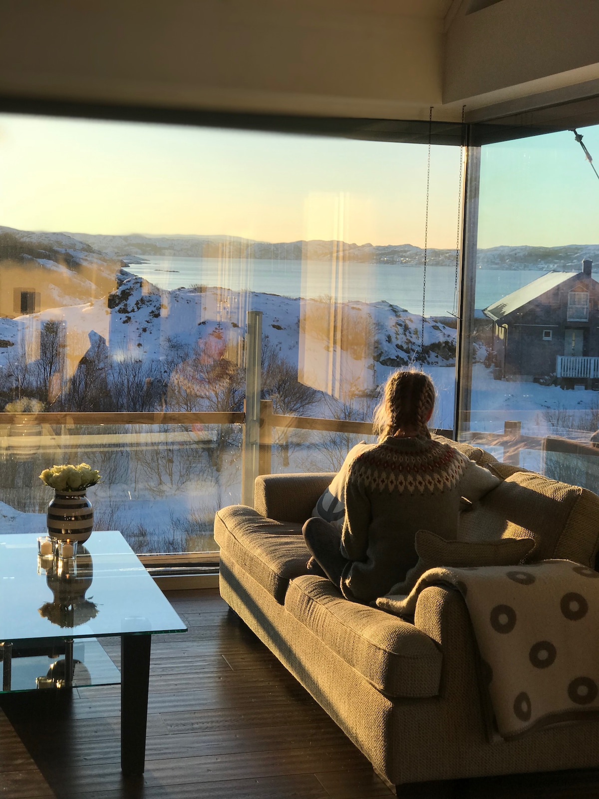 A modern living area features large glass windows, offering a scenic view of the snowy landscape and water beyond. A comfortable sofa is positioned near the window, with a small table displaying a vase of flowers. Soft natural light fills the space.