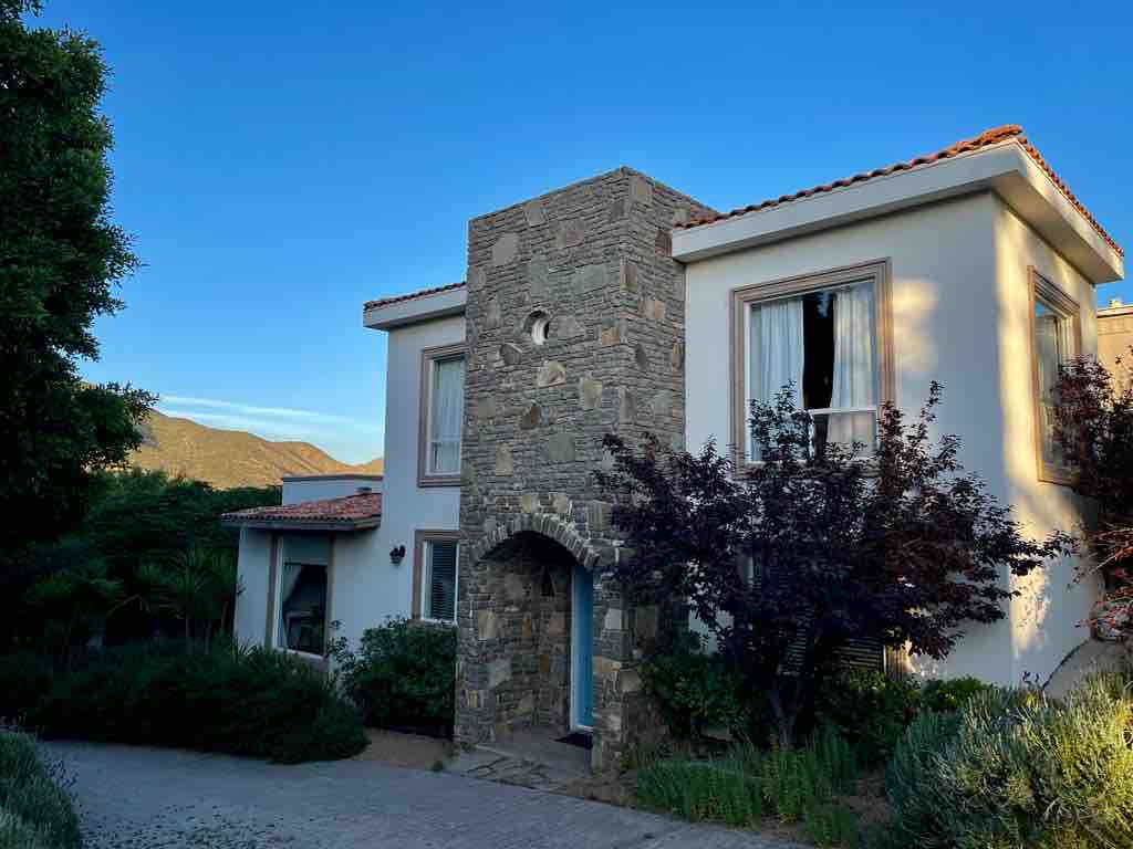 A two-story building features a stone facade and traditional tiled roof. Large windows provide natural light, while a welcoming entrance is framed by greenery. The surrounding landscape includes distant hills, enhancing the peaceful setting.