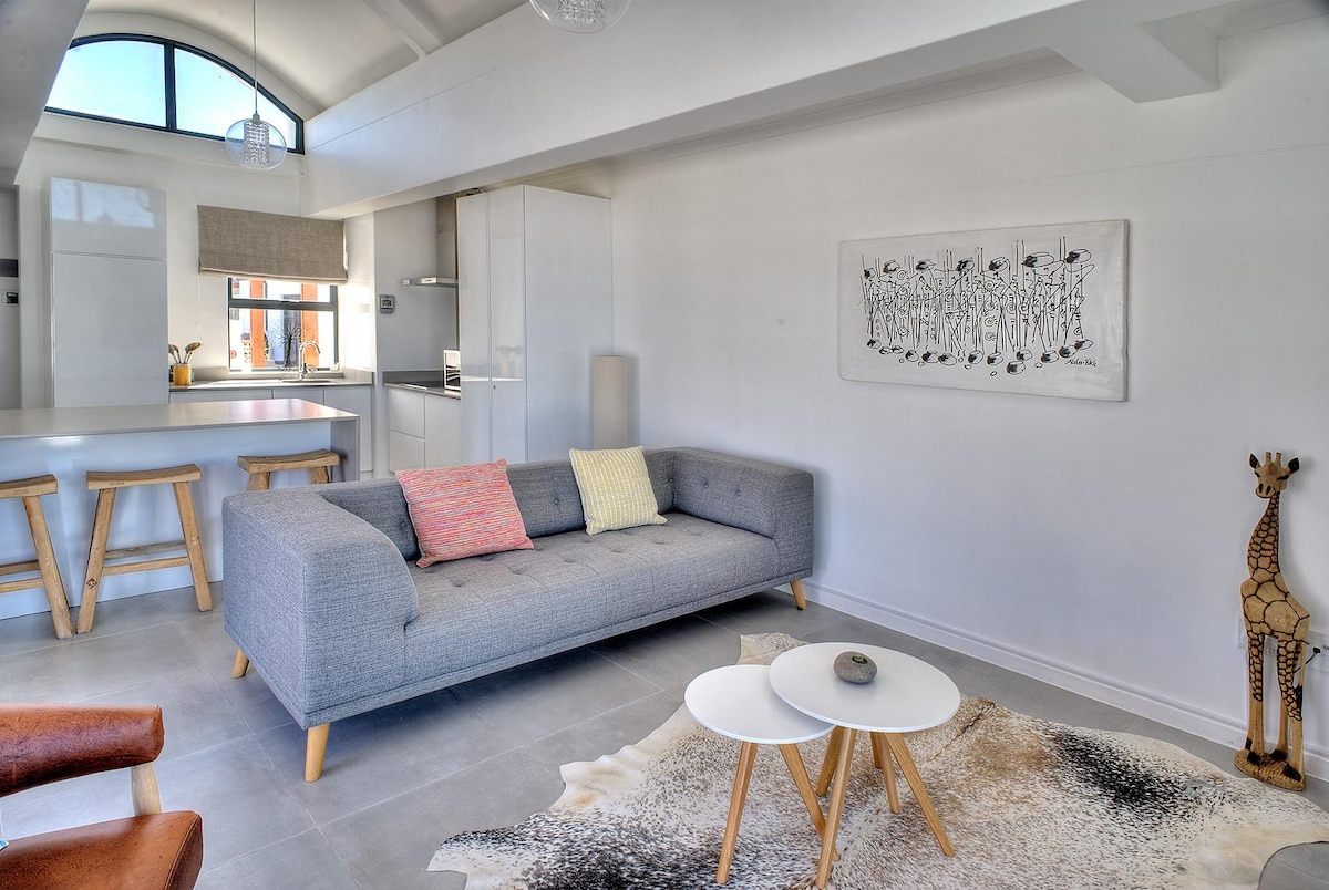 A modern living space features a light grey sofa with colorful cushions, paired with a white coffee table atop a textured rug. A minimalist kitchen is visible in the background, along with wooden stools. Artistic wall decor enhances the inviting environment.