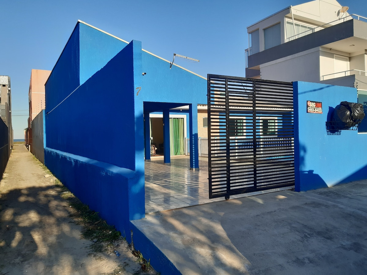 A vibrant blue exterior of a single-story house is framed by modern fencing. The entrance is inviting, revealing a spacious outdoor area with tiled flooring. Light filters through large windows, suggesting an airy atmosphere inside. Nearby structures are visible in the background.