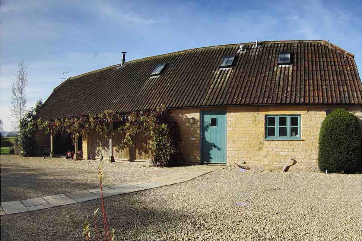 A detached cottage with a stone facade is shown, featuring a sloping roof and charming blue front door. Lush greenery frames the porch area, and a pathway leads up to the entrance. The surrounding grounds are filled with gravel and offer open sky views.