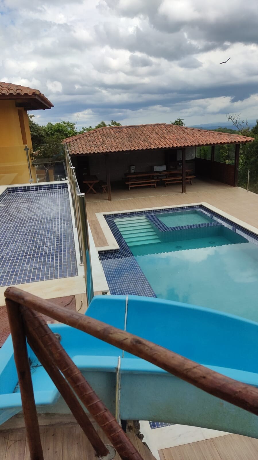 An outdoor pool area is visible, featuring a spacious swimming pool with attached lounge area. A waterslide descends into the pool. A covered gazebo with seating is nearby, surrounded by landscaped grounds under a partially cloudy sky.