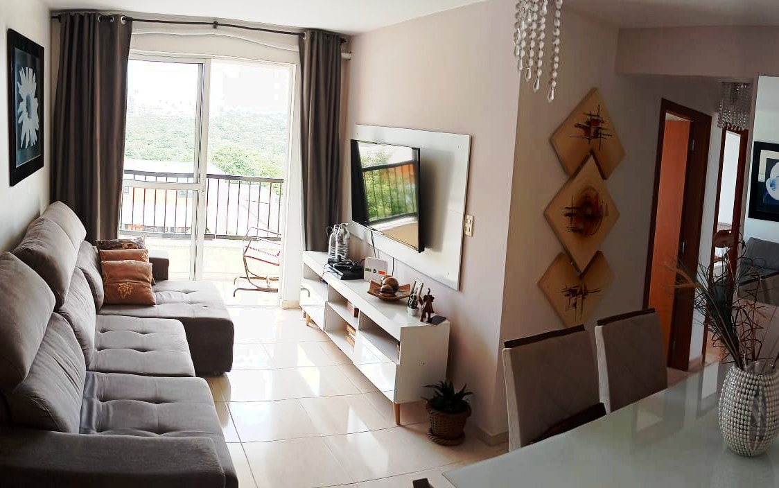 A spacious living area features a light-colored sectional sofa accompanied by decorative cushions. A flat-screen TV is mounted on the wall opposite a modern media console. Large windows allow natural light to fill the room, offering views of the surrounding landscape.