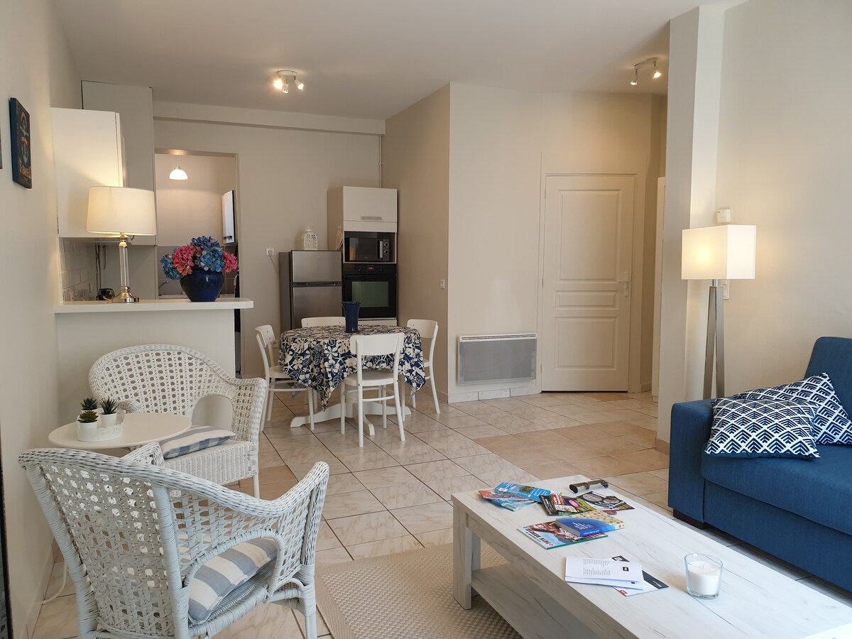 A bright living area features a comfortable sofa, a coffee table, and a dining set with four chairs. The open kitchen with modern appliances, including a refrigerator and microwave, is visible in the background. Light-colored tiles and soft lighting contribute to a welcoming environment.