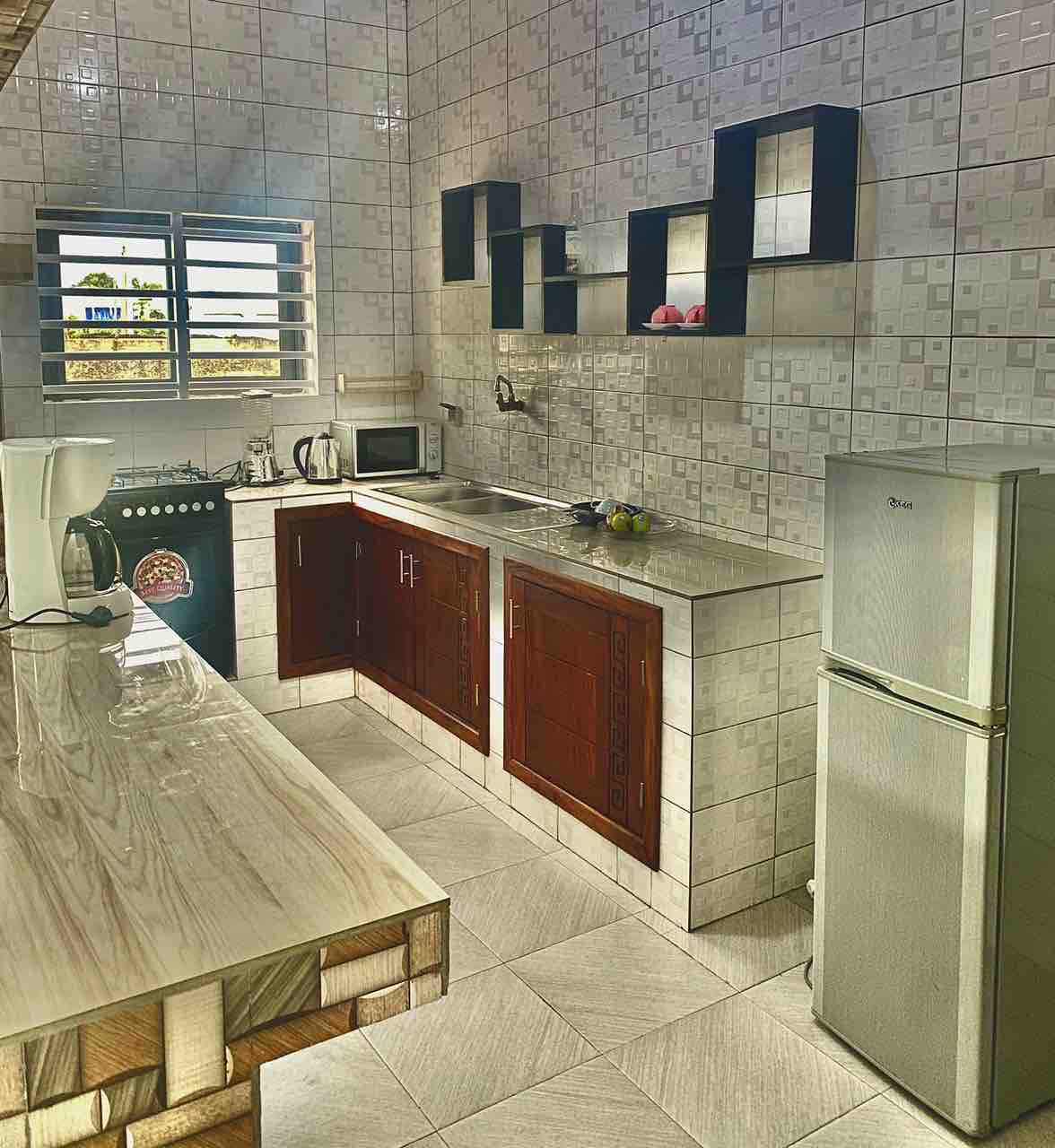 The kitchen features modern cabinetry with a stylish countertop and built-in shelving. A refrigerator and microwave are positioned for convenience. Natural light enters through a window, illuminating the space, which includes kitchen appliances such as a coffee maker and stovetop.