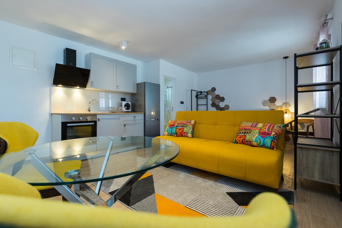 A bright living area features a yellow sofa adorned with colorful cushions, set against a stylish geometric rug. A glass dining table reflects gentle lighting, while a well-equipped kitchen with modern appliances is situated in the background, providing a functional and inviting space.