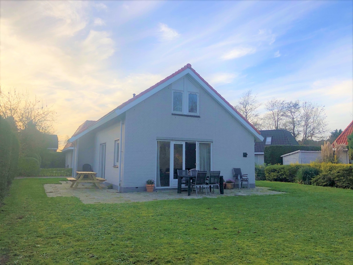 A spacious villa is set within a lush garden, with a large lawn area and a patio featuring seating and a dining table. Large windows offer views into the home while the peaceful surroundings provide a sense of privacy.
