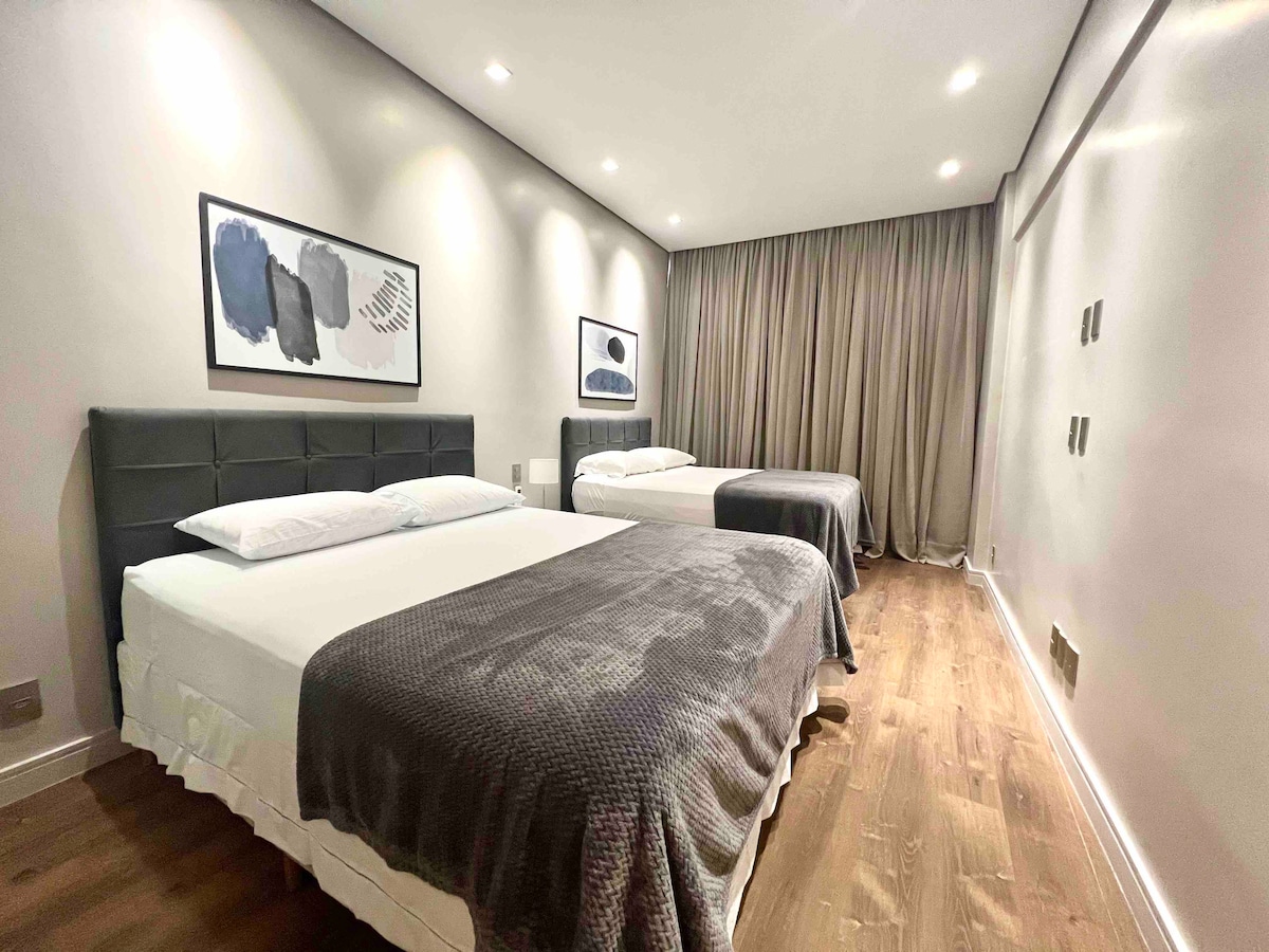 Two spacious beds are arranged in a well-lit bedroom, featuring soft gray blankets and large windows covered by flowing curtains. Modern artwork adorns the wall above the beds, contributing to a serene ambiance.