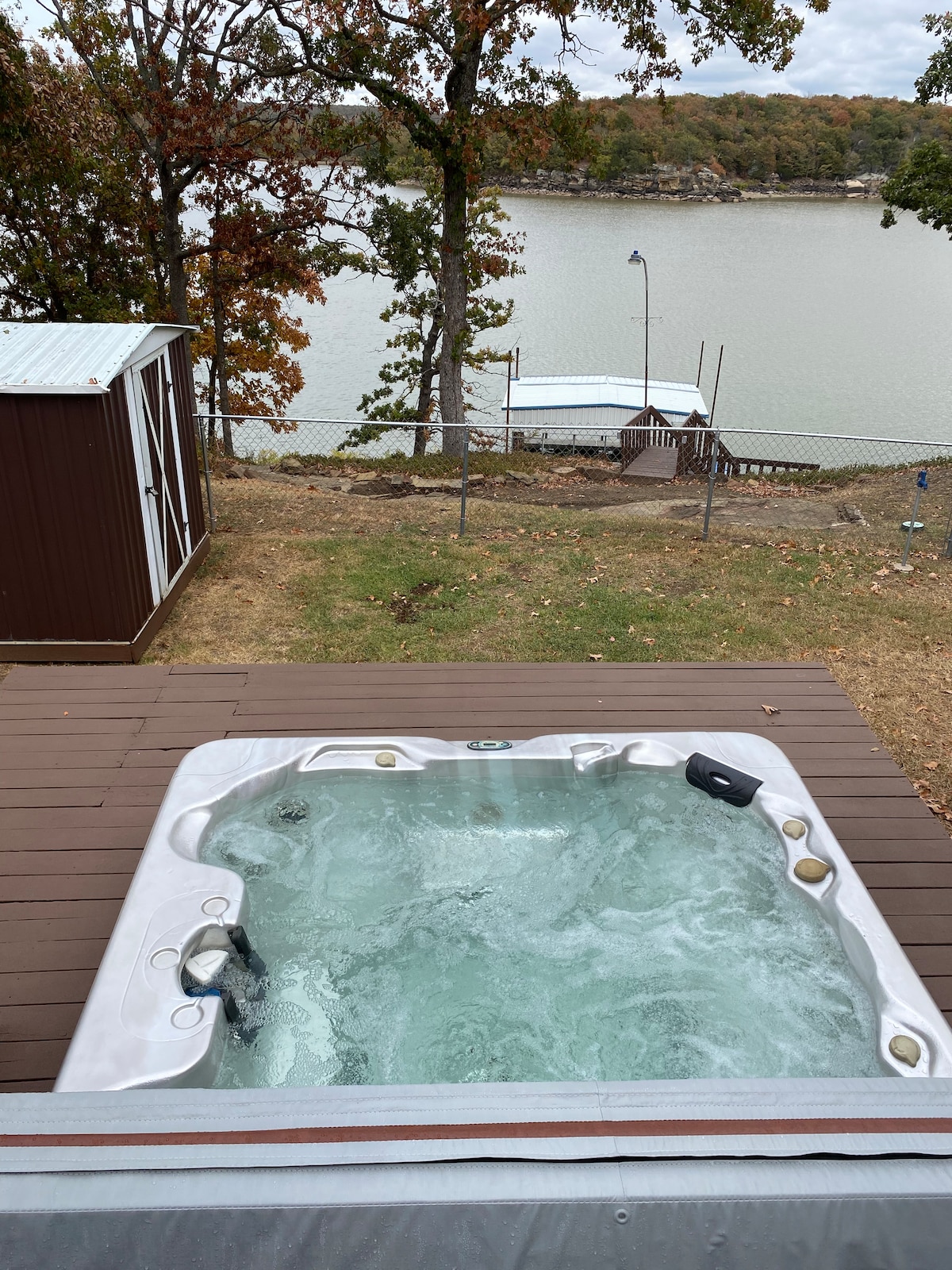 A hot tub is situated on a wooden deck, overlooking a serene lake. The bubbling water reflects the surrounding autumn foliage, while a private dock is visible in the background, adding to the tranquil lakeside setting.