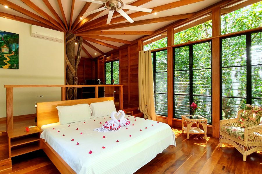 A spacious bedroom features a king-size bed decorated with rose petals, surrounded by floor-to-ceiling windows that bring in natural light. A cozy chair is positioned nearby, while wooden beams create an inviting ceiling design, integrating the room with the lush greenery outside.