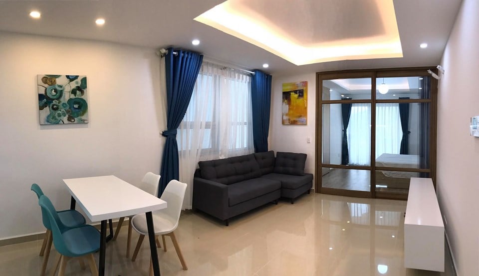 A modern living area is presented with a plush gray sofa, a white dining table surrounded by colorful chairs, and vibrant artwork adorning the walls. Large windows are framed by blue curtains, allowing natural light to enhance the space. A glass partition leads to the bedroom beyond.