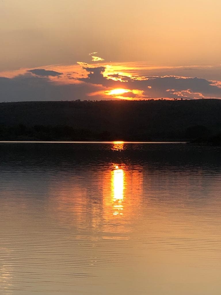 The image captures a serene sunset over a body of water, with warm orange and yellow hues reflecting on the calm surface. Silhouetted hills provide a gentle backdrop, while cloud formations add depth to the evening sky.