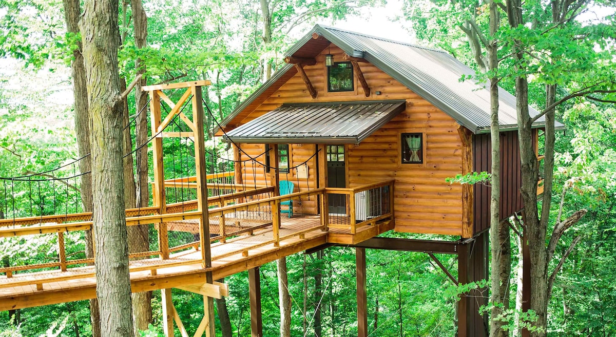 A charming treehouse is nestled among lush greenery, accessed via a wooden walkway. The exterior features natural wood siding and a metal roof, complemented by a balcony with outdoor seating that invites relaxation amidst the surrounding trees.
