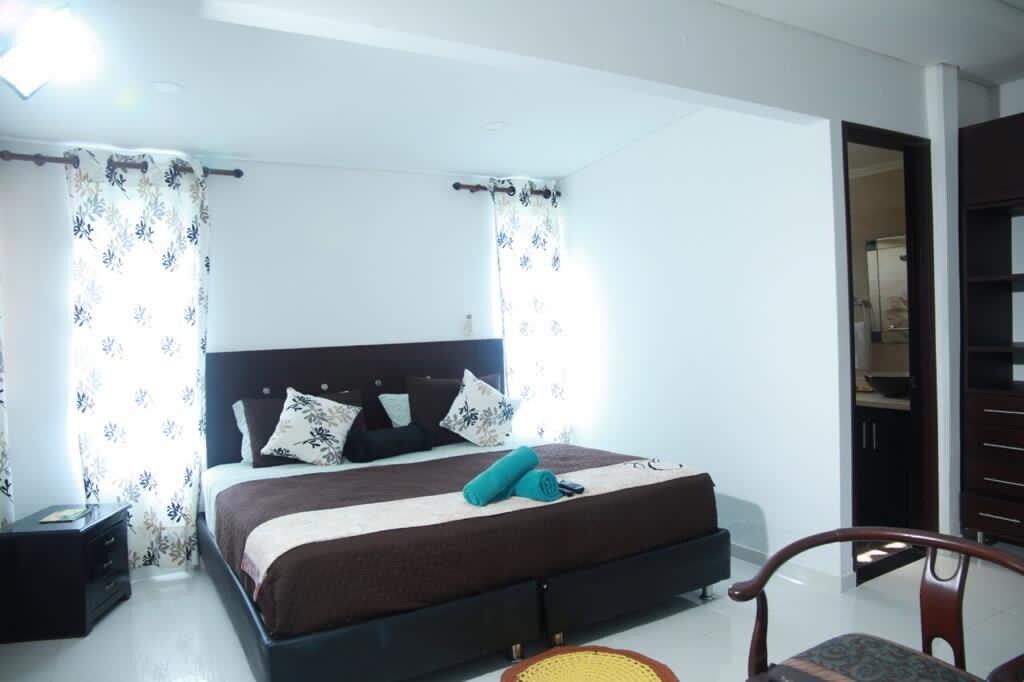 A spacious bedroom features a large bed with decorative pillows and a folded towel placed at the foot. Natural light enters through two window curtains, complemented by a dark cabinet and a small round table with chairs nearby. An entrance to a bathroom is visible in the background.