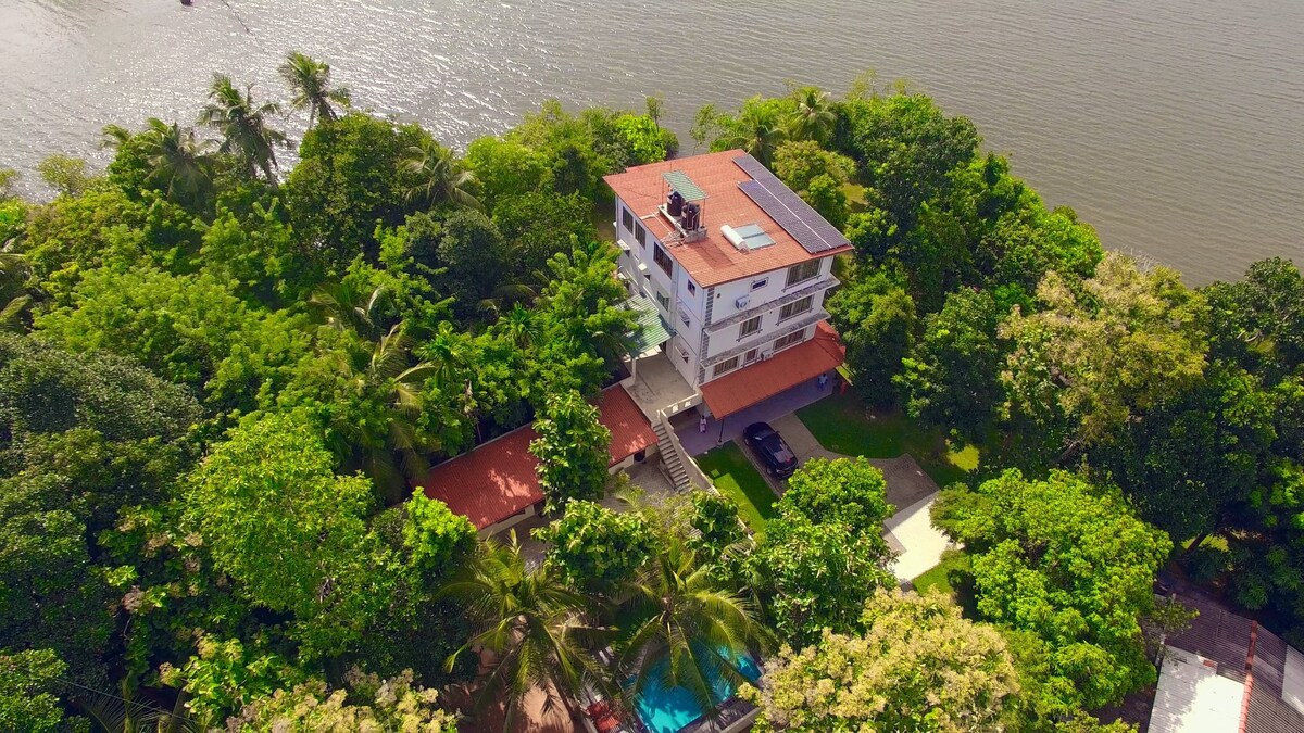An aerial view captures the three-story Holiday Home surrounded by lush greenery and mature trees. The property is positioned near the Madu River, with a pool visible in the garden area. The terracotta roof complements the vibrant natural landscape, creating a serene retreat atmosphere.