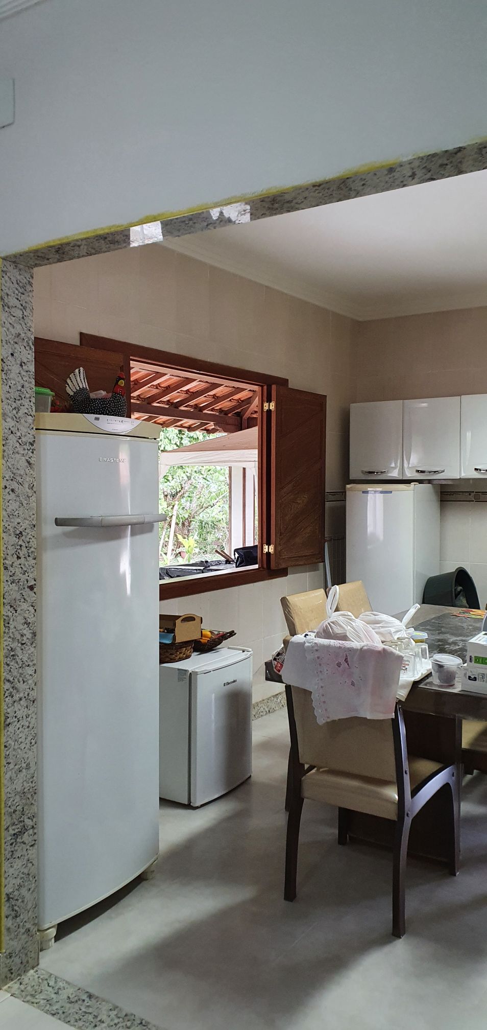 A spacious kitchen area is highlighted with a tall refrigerator and a smaller freezer nearby. A wooden dining table, surrounded by chairs, shows various kitchen utensils. Natural light enters through a wide window with wooden shutters, revealing greenery outside.