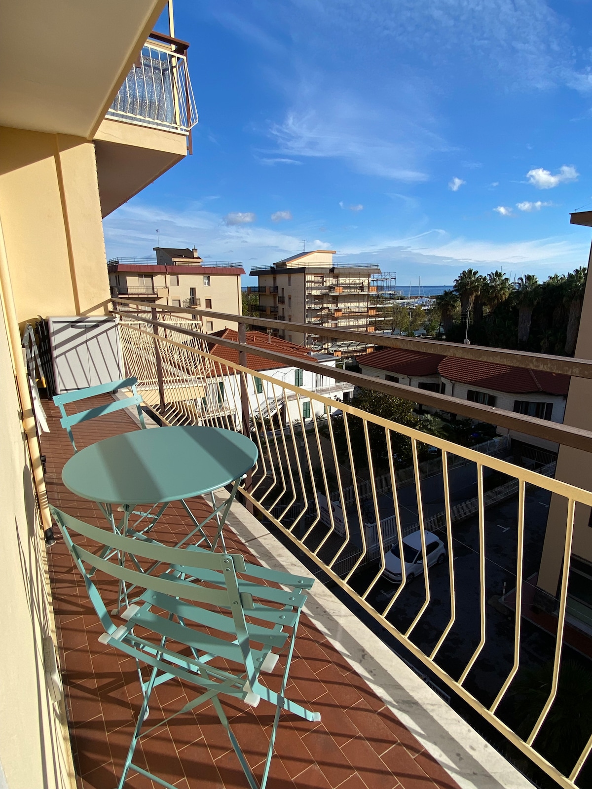 A balcony features a small round table and two chairs, both in a soft blue hue. The railing offers an unobstructed view of the surrounding buildings and distant sea under a clear sky. Sunlight creates a bright ambiance on the outdoor space.