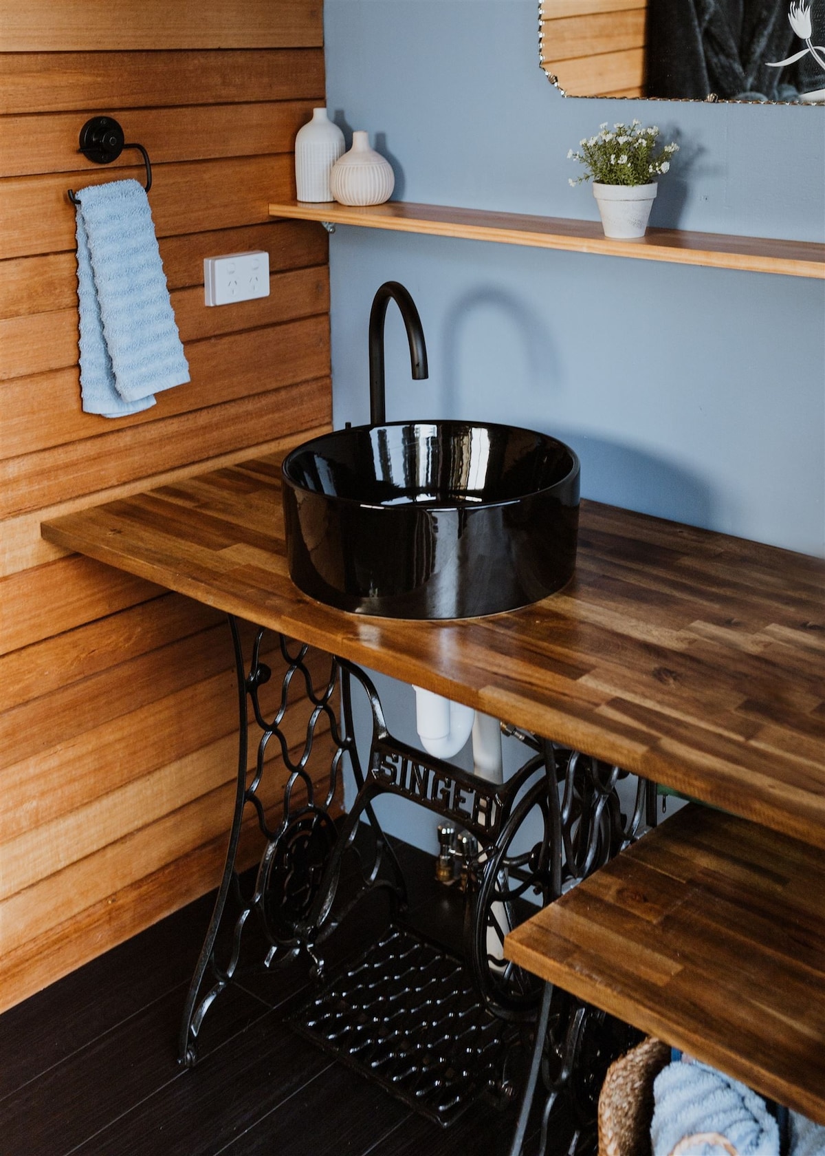A stylish bathroom sink area features a round black vessel sink resting on a wooden countertop. The natural wood paneling complements the modern faucet, and subtle decor items, including a small vase with greenery, add a touch of charm.