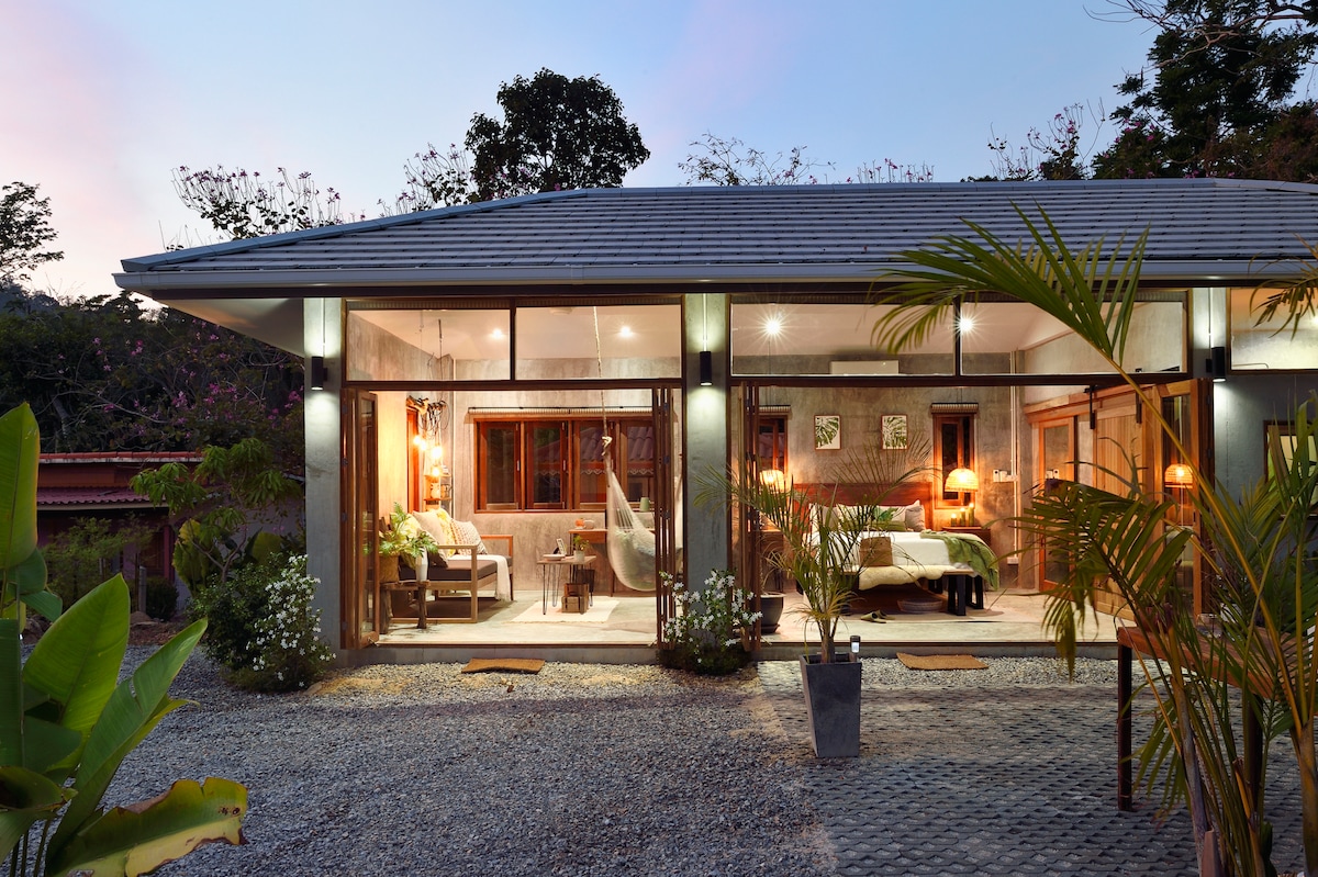The villa's exterior is framed by lush greenery and a gravel pathway. Large glass doors reveal a bright, open space where tasteful decor and cozy seating invite relaxation. The tranquil setting is complemented by ambient lighting as the sun sets in the background.