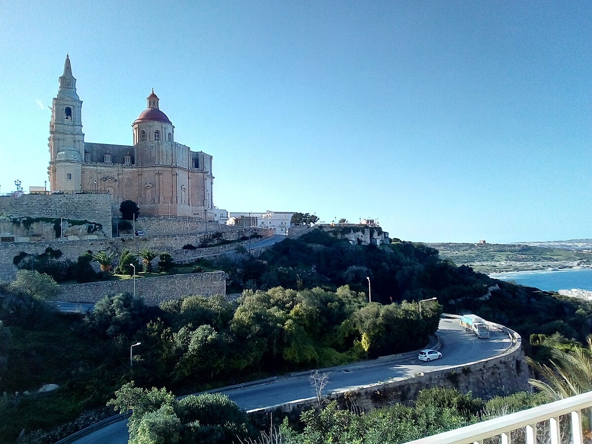 A serene landscape captures a church situated on a hilltop, surrounded by lush greenery and distant sea views. The smooth road winds down the hillside, with a vehicle visible. Clear skies provide a vibrant blue backdrop, enhancing the scene's tranquility.