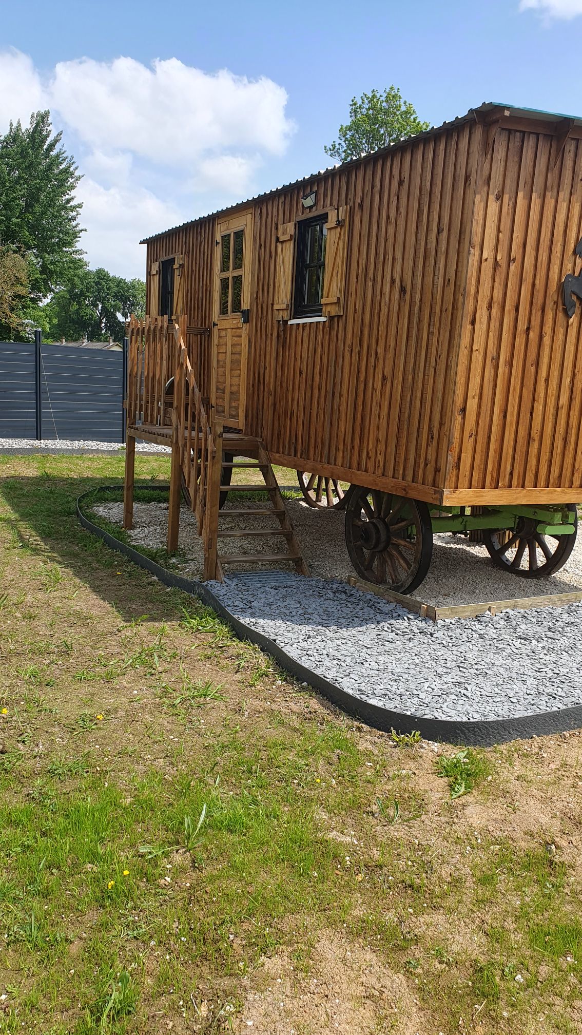 A charming wooden caravan is presented atop sturdy wheels, featuring a staircase leading to the entrance. The exterior is framed by lush greenery with a gravel pathway bordering the structure. Sunlight casts a warm glow on the natural wood finish.