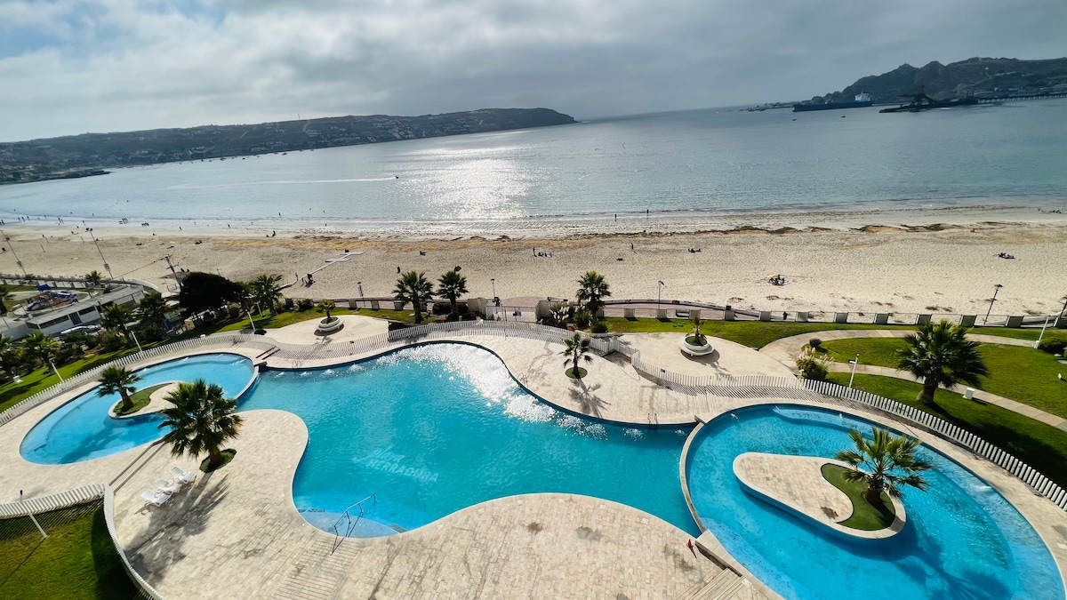 A spacious outdoor area features a large, winding pool surrounded by manicured gardens. Palm trees provide shade, and the sandy beach is visible in the background, leading to a calm ocean under a cloudy sky.
