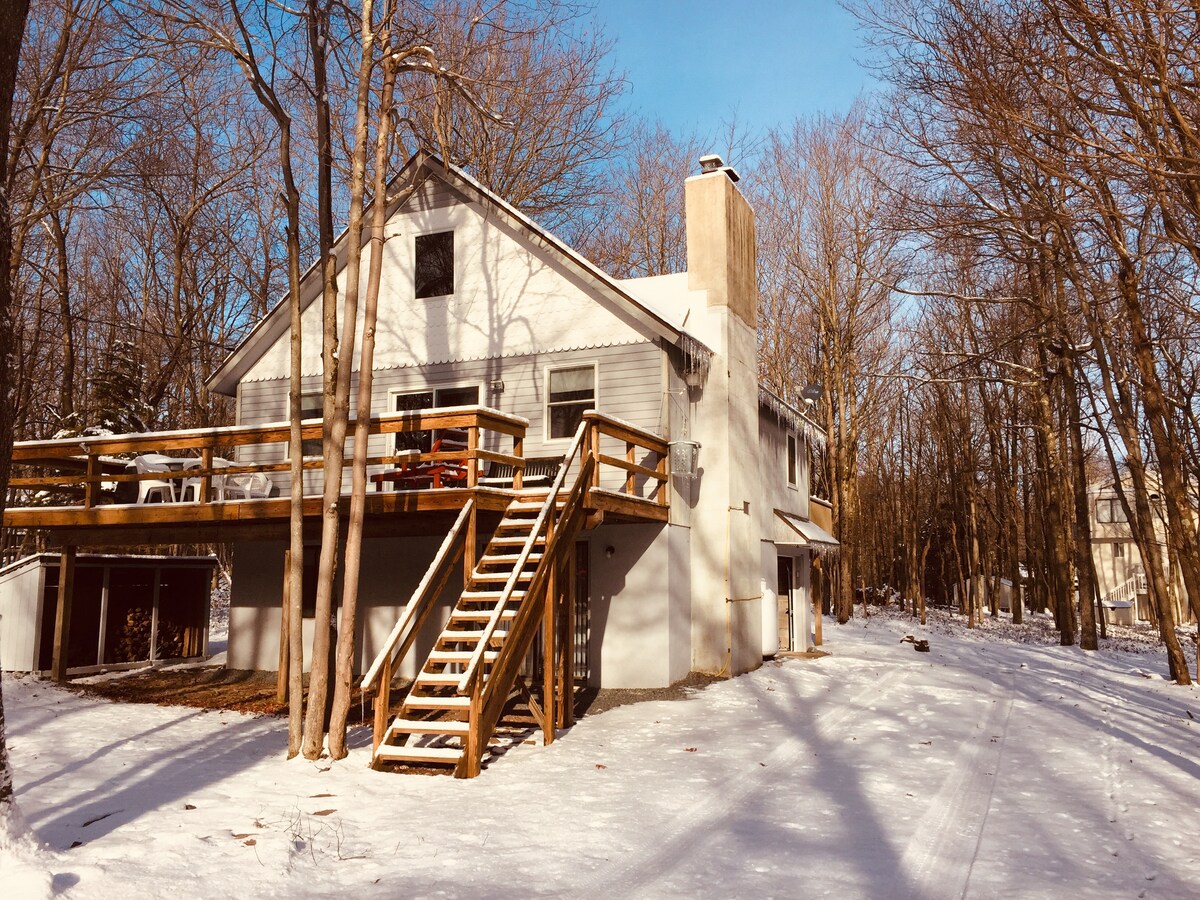 A cozy chalet is surrounded by snow-covered trees, featuring a large wooden deck with stairs leading to the entrance. The exterior showcases a light-colored façade and a chimney, reflecting a tranquil winter atmosphere.