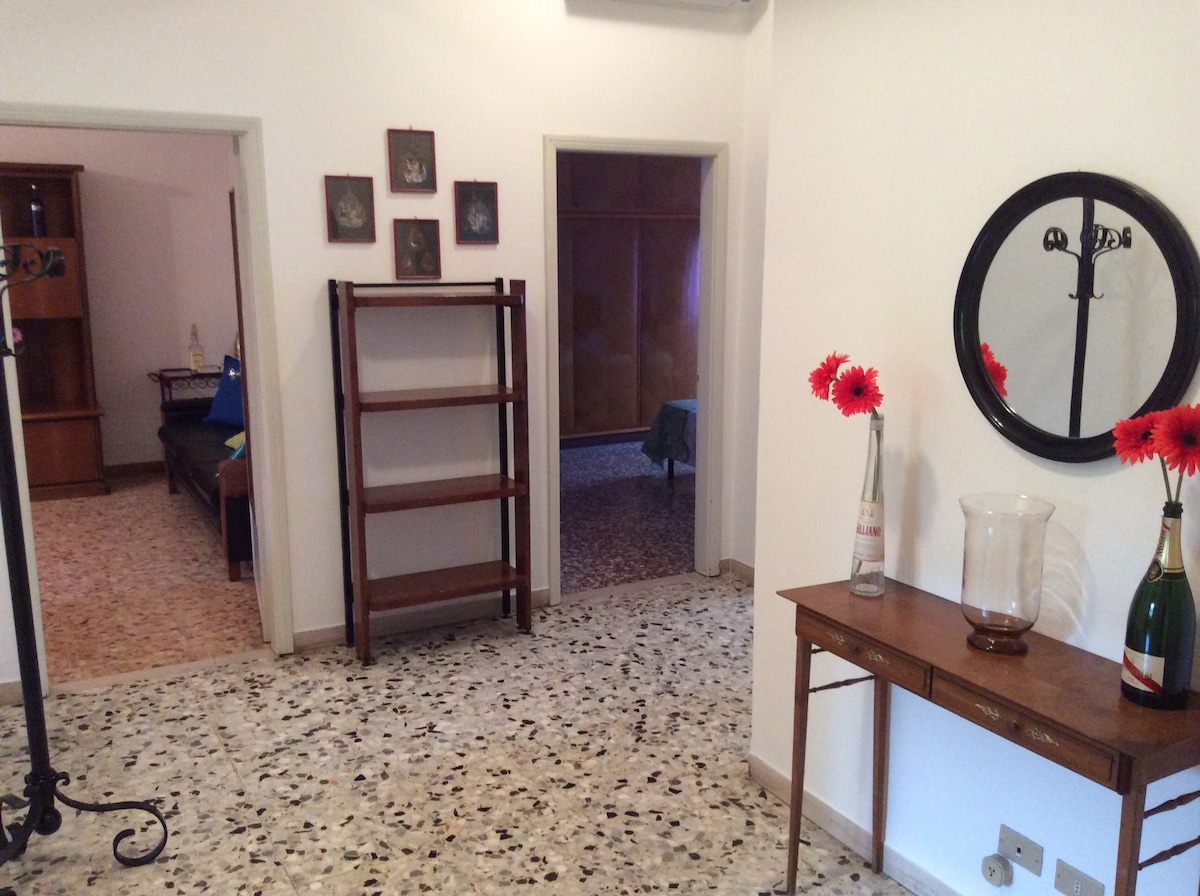 An open hallway is visible, featuring a decorative table with a vase and flowers. A shelf stands against the wall, and a round mirror is mounted nearby. The flooring is a mix of light and dark tiles, contributing to the space's overall brightness.